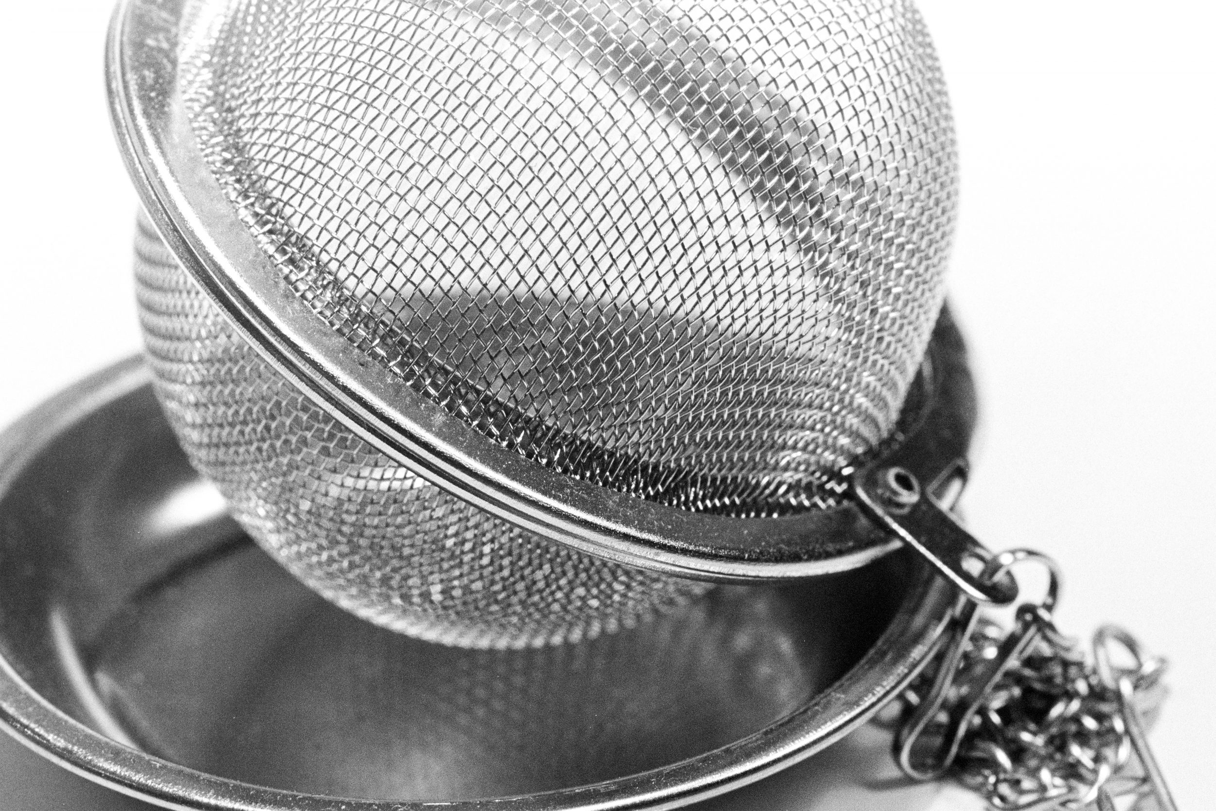 Tea Strainer - Tea Infuser - A close-up shot of a silver metal tea strainer, also known as a tea infuser. The fine mesh design of the infuser is clearly visible, with intricate details highlighting the texture of the metal. The strainer's sleek and compact form contrasts with the soft grayscale tones of the film on which it was taken, creating a timeless and elegant look. The delicate mesh and silver finish emphasise the practical yet refined nature of this kitchen tool.